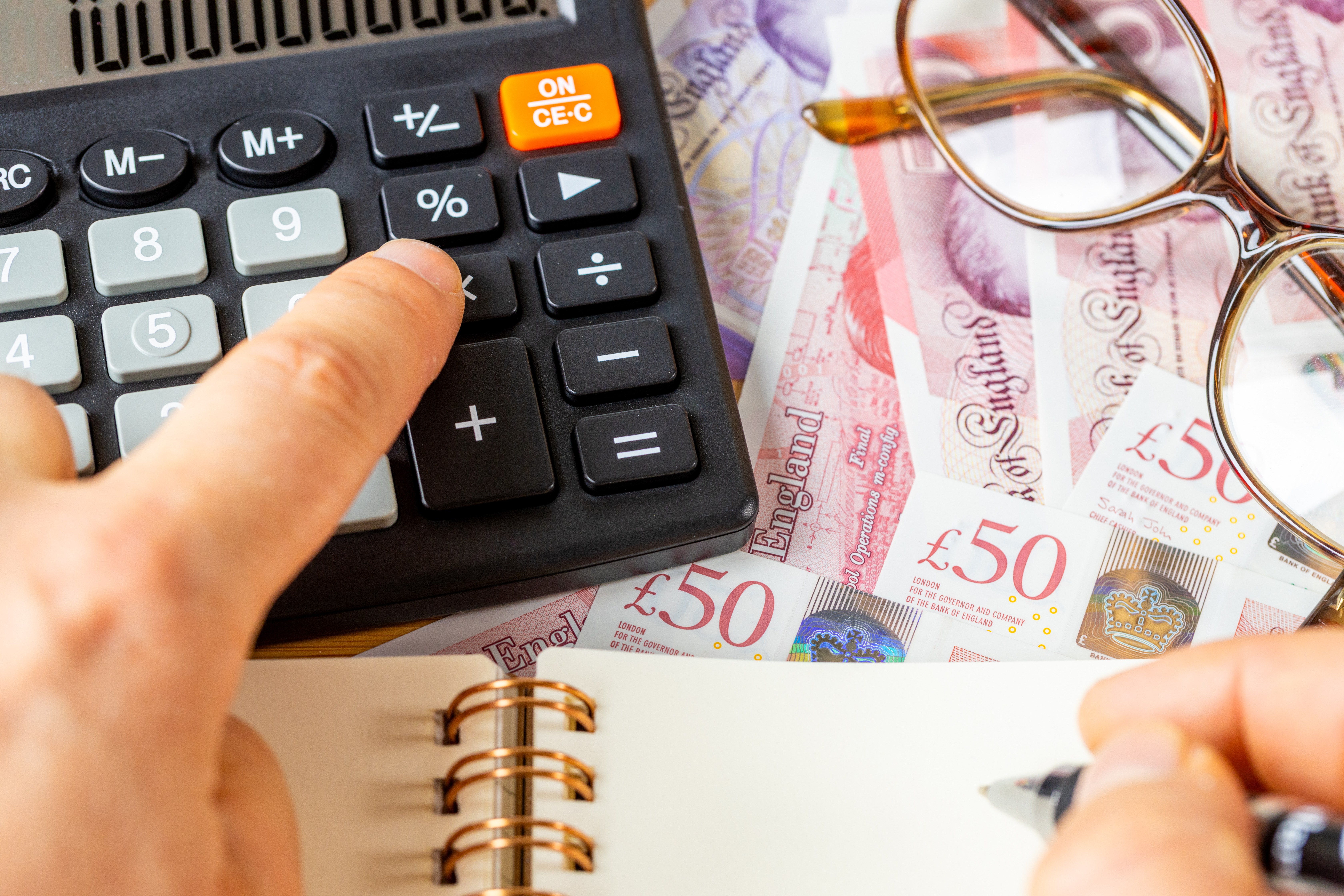 Person typing into calculator on desk covered in money, glasses and notebook