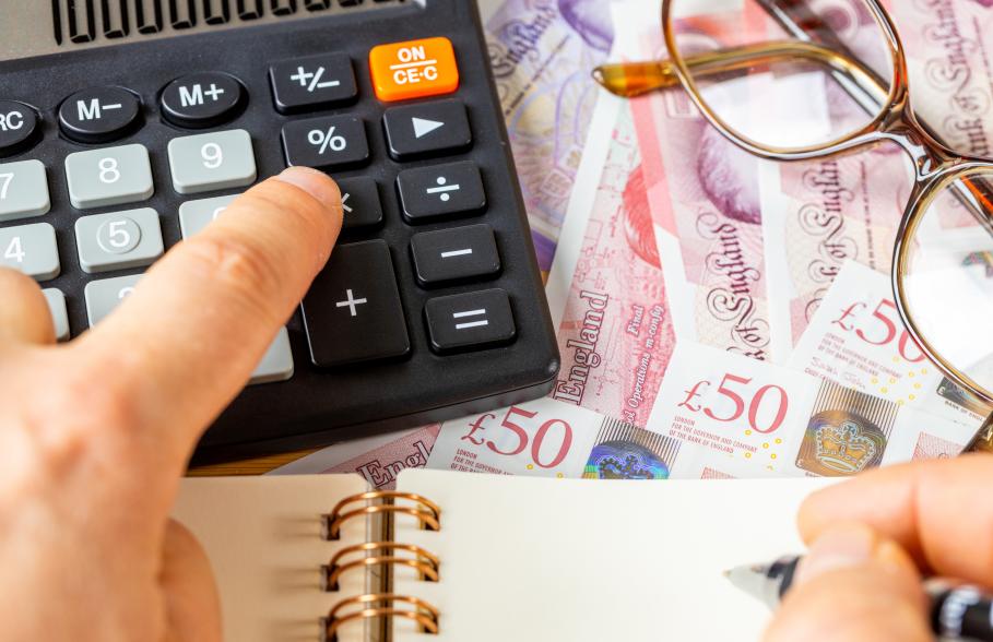 Person typing into calculator on desk covered in money, glasses and notebook