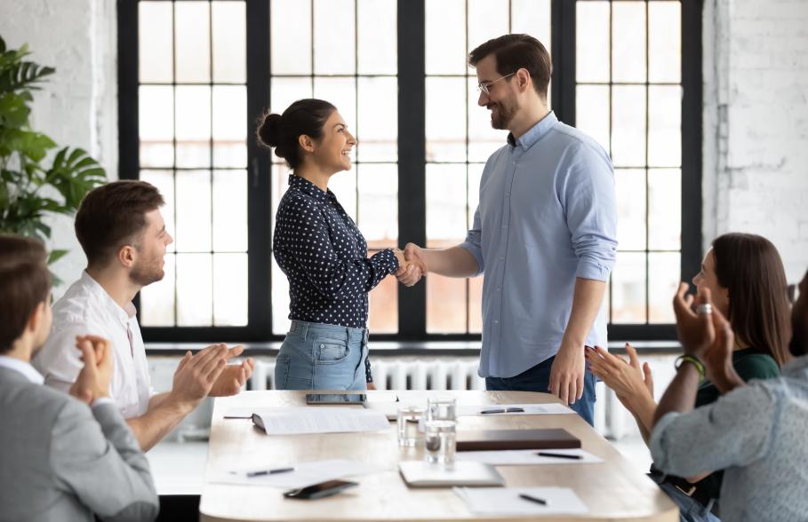 A person gives a handshake to another person whilst others in the room applaud