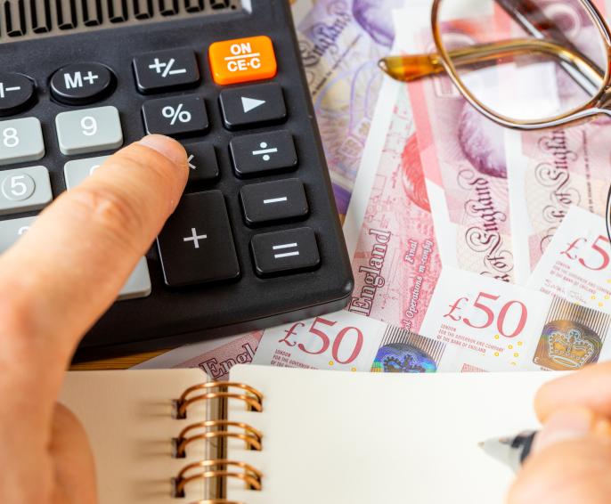 Person typing into calculator on desk covered in money, glasses and notebook